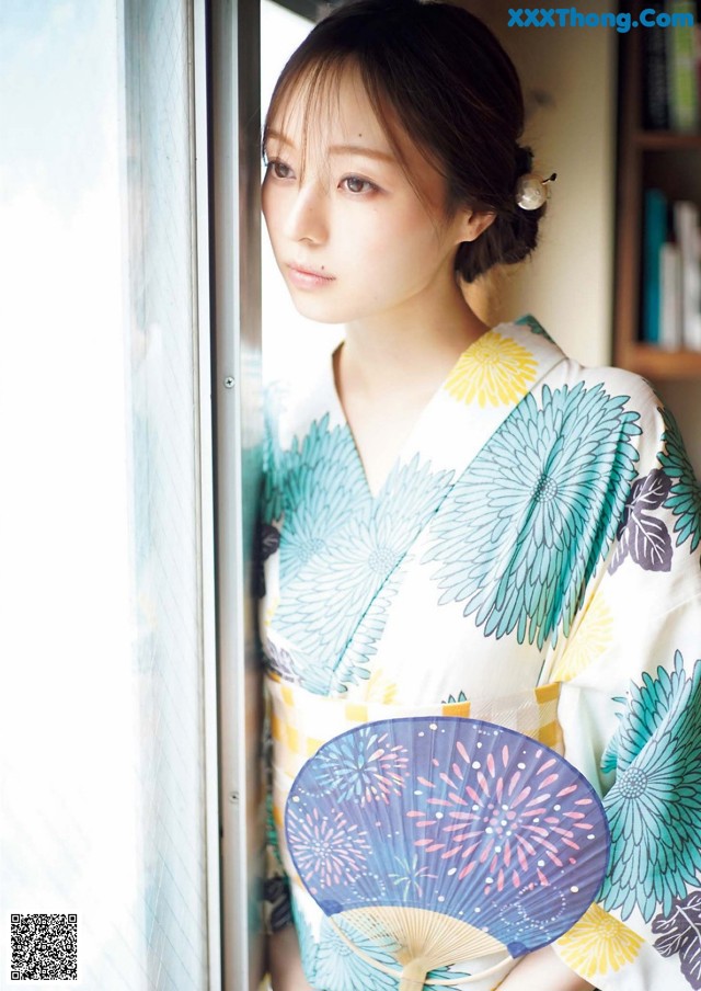 A woman in a kimono holding a fan by a window.